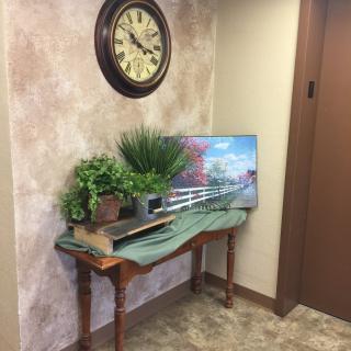 A hallway corner features a table with decorative plants, a picture frame, and an ornate wall clock.
