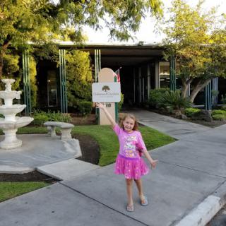A cheerful child in a pink dress holding up a sign near a fountain on a sunny day.