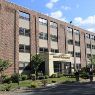 A multi-story brick building with several windows and a landscaped area in front, under a blue sky with clouds.