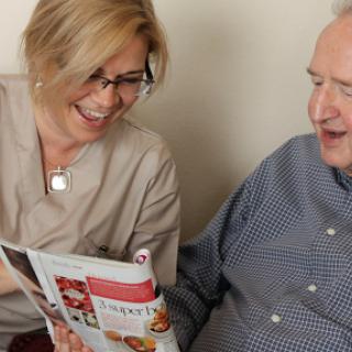 A caregiver and an elderly man share a joyful moment, looking at a magazine together.