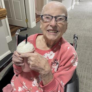 An elderly woman in a pink sweater enjoys a milkshake while smiling in her wheelchair.
