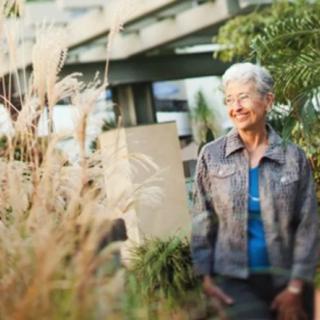 An elderly woman smiling while standing in a garden surrounded by plants.