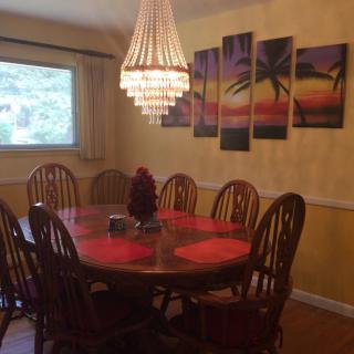 A cozy dining room featuring a wooden dining table with red placemats, surrounded by chairs and illuminated by a chandelier. The wall is adorned with a colorful piece of artwork depicting palm trees.