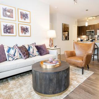 A modern living room featuring a white sofa with patterned pillows, a brown leather armchair, and a round coffee table, complemented by framed artwork on the wall.