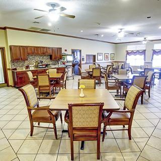 A spacious dining area with neatly arranged tables and chairs, featuring a tiled floor and kitchen facilities in the background.