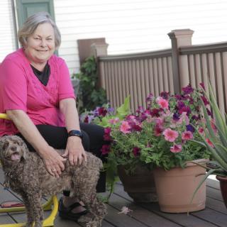 A woman sitting on a porch with a dog beside her, surrounded by potted flowers.