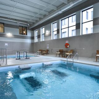 This image shows a spacious indoor swimming pool with a relaxation area featuring tables and chairs, well-lit by large windows.