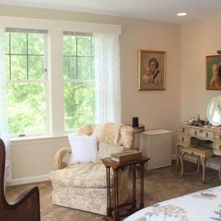 A cozy living room with vintage decor, featuring an armchair, artwork on the walls, and a bright window view.