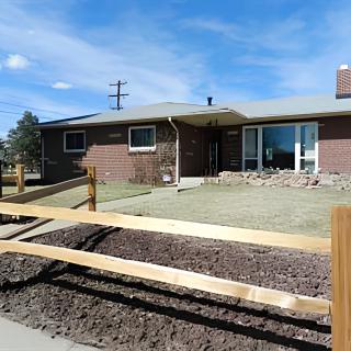 A single-story brick house with a front lawn and wooden fence, set against a clear blue sky.