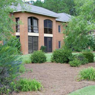 A brick building with large windows surrounded by lush greenery and landscaped bushes.