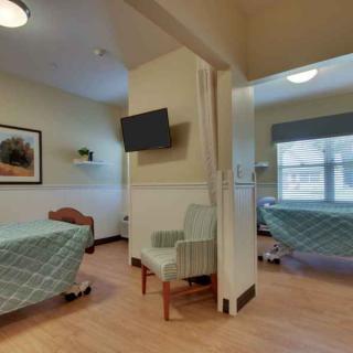 A hospital room with patient beds separated by curtains, featuring a television and chairs.