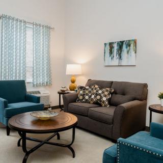A cozy living room featuring a comfortable sofa, two blue armchairs, and a round coffee table. The space is accented with a lamp and wall art.