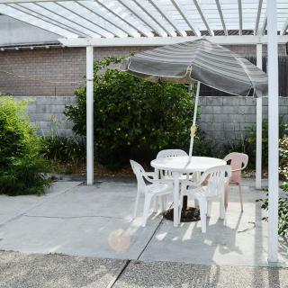 A backyard patio featuring white plastic chairs and a table under a pergola, with an umbrella for shade.