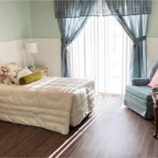 A cozy bedroom with a neatly made bed, an armchair by the window, and sheer curtains letting in natural light.