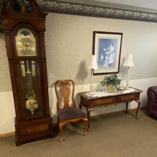 A cozy room corner featuring a grandfather clock, a wooden chair, and a table topped with decorative items beneath wall art.