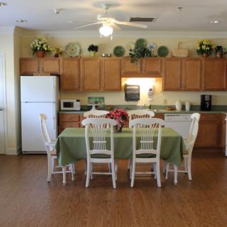 A cozy kitchen and dining area featuring wooden cabinets, a refrigerator, a microwave, and a table set with chairs. The space is decorated with plants and flowers.