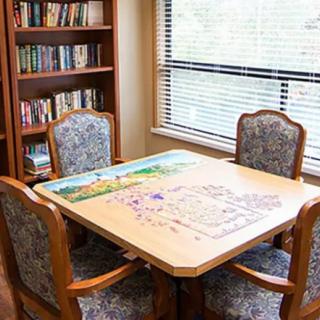 A cozy indoor setting with a table and chairs, featuring a partially completed puzzle, next to a bookshelf by the window.