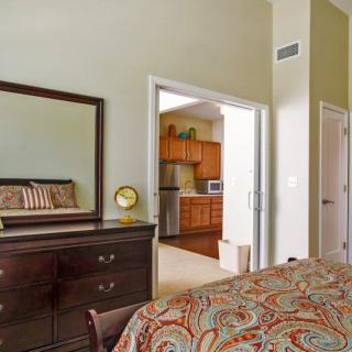A cozy bedroom with a view into the kitchen, featuring a dresser with a mirror and patterned bedspread.