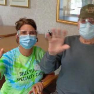 Two people wearing masks and glasses are sitting indoors, waving at the camera.