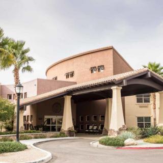 The image shows the entrance of a modern building surrounded by palm trees under a cloudy sky.
