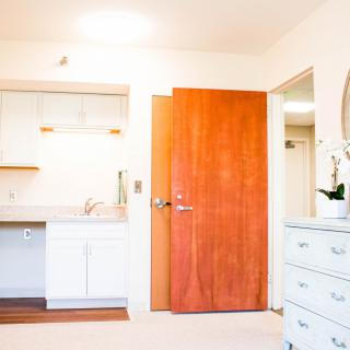 A bright minimalist apartment interior featuring a small kitchen area, white walls, and wooden door.