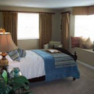 A cozy bedroom featuring a bed with blue and white bedding, two windows with curtains, and a sitting area by the window.