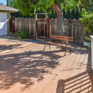 A sunny backyard with a wooden deck, featuring a bench and tree surrounded by a fence.