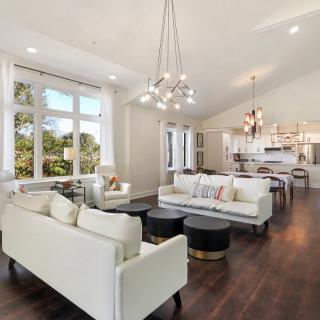 A modern open-concept living room with a view into the kitchen, featuring white sofas, hardwood floors, and elegant chandeliers.