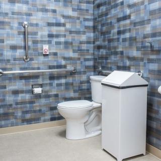 An accessible bathroom with a toilet, grab bars, and a trash can against a tiled wall.