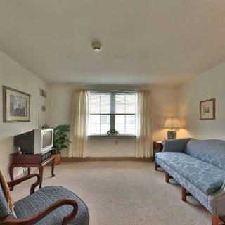 A cozy living room with a sofa, armchair, and television, softly lit by a lamp near the window.