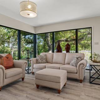 A cozy living room with a sofa, armchair, and ottoman set against large windows offering a view of greenery.