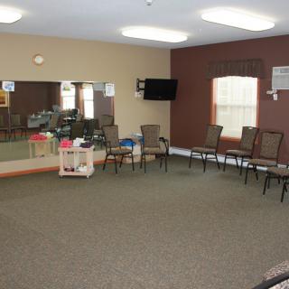 A spacious room with chairs arranged in a semi-circle, featuring mirrors, a wall-mounted television, and windows.