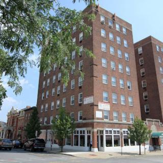A view of a multi-story brick building in an urban setting, surrounded by trees and other structures.
