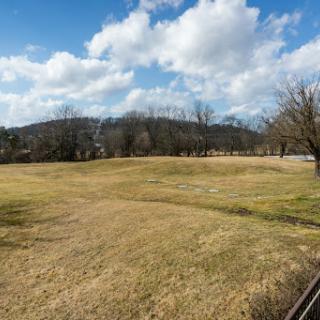 A serene view of an open field under a partly cloudy sky, surrounded by trees and distant hills.