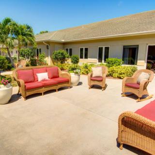 A sunny patio with wicker furniture and red cushions, surrounded by lush greenery.