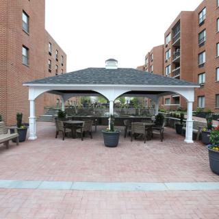 Outdoor gazebo area with tables, benches, and potted plants surrounded by brick buildings.