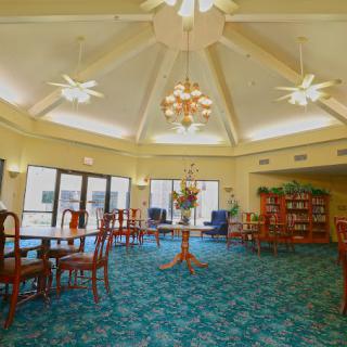 A spacious room with a high ceiling, featuring tables and chairs on a patterned carpet, surrounded by large windows and bookshelves.