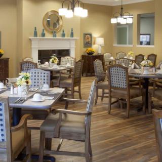 A cozy dining room with neatly arranged tables and chairs, featuring a warm fireplace and elegant chandeliers.