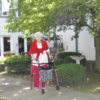 An elderly woman using a walker outside a house with trees in the background.