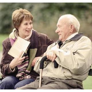 A woman and an elderly man are sitting on a bench in a park, engaged in a friendly conversation. The woman is holding a book, and both are smiling.