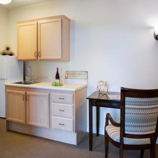 A compact kitchenette with wooden cabinets, a small desk, and a chair in a cozy indoor setting.