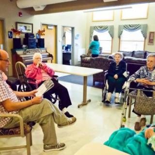 A group of elderly people sitting in a nursing home common area, some using wheelchairs.
