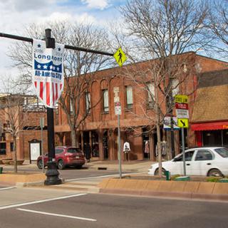 A view of a downtown street with brick buildings, cars parked along the road, and a crosswalk in the foreground.