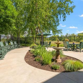 A tranquil outdoor garden with a patio, fountain, and seating under trees on a sunny day.