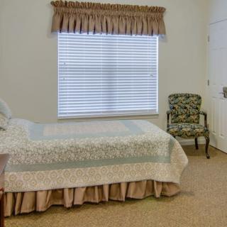 A cozy bedroom with a single bed covered by a quilt, a window with blinds and curtains, and a patterned chair in the corner.