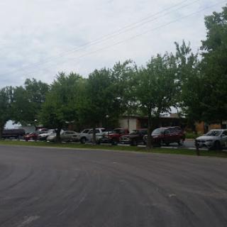A view of a street lined with parked vehicles and lush green trees under a cloudy sky.