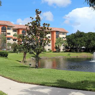 A serene view of an apartment complex by a tranquil pond, surrounded by lush greenery and a clear blue sky.