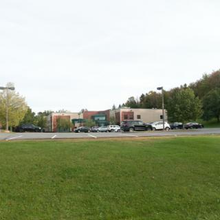 A view of a commercial building with a parking lot surrounded by grass and trees under a cloudy sky.
