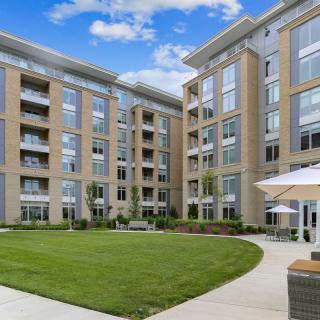 A modern apartment building with a well-maintained courtyard and green lawn, featuring outdoor seating and umbrellas.