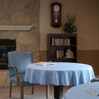 A cozy room with a table covered in a blue cloth, surrounded by chairs. A wooden bookshelf and a wall clock add to the decor, while a stone fireplace provides warmth.